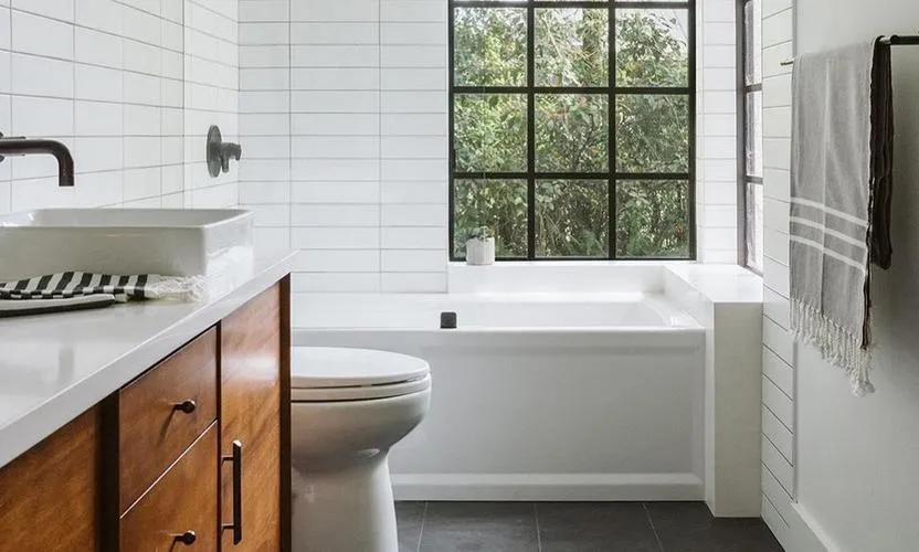 A modern bathroom interior featuring white tiles, a black countertop, wooden cabinets, and stainless steel fixtures. The space includes a toilet, sink with window curtains, and a tiled shower area.